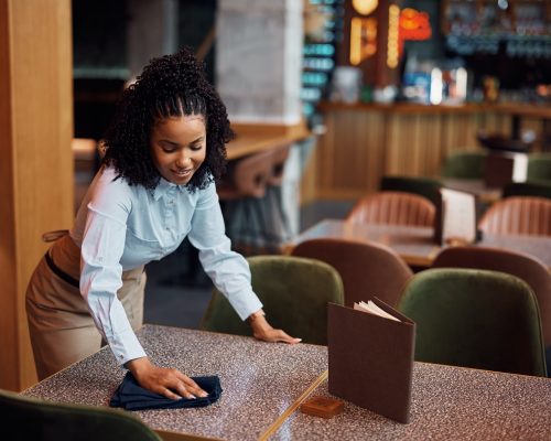 Happy African American waitress cleaning tables in a cafe.